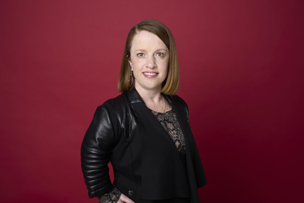 Photo of Rebecca Cokley wearing a leather blazer over a patterned blouse, standing against a red background.