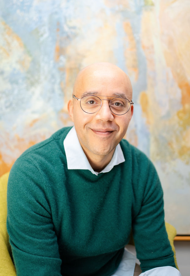 Amiddle-aged, light skinned, Black male wearing gold wire rimmed circular classes, a blue collared shirt, and a green sweater, seated in front of a pastel blue, orange, and white painting.