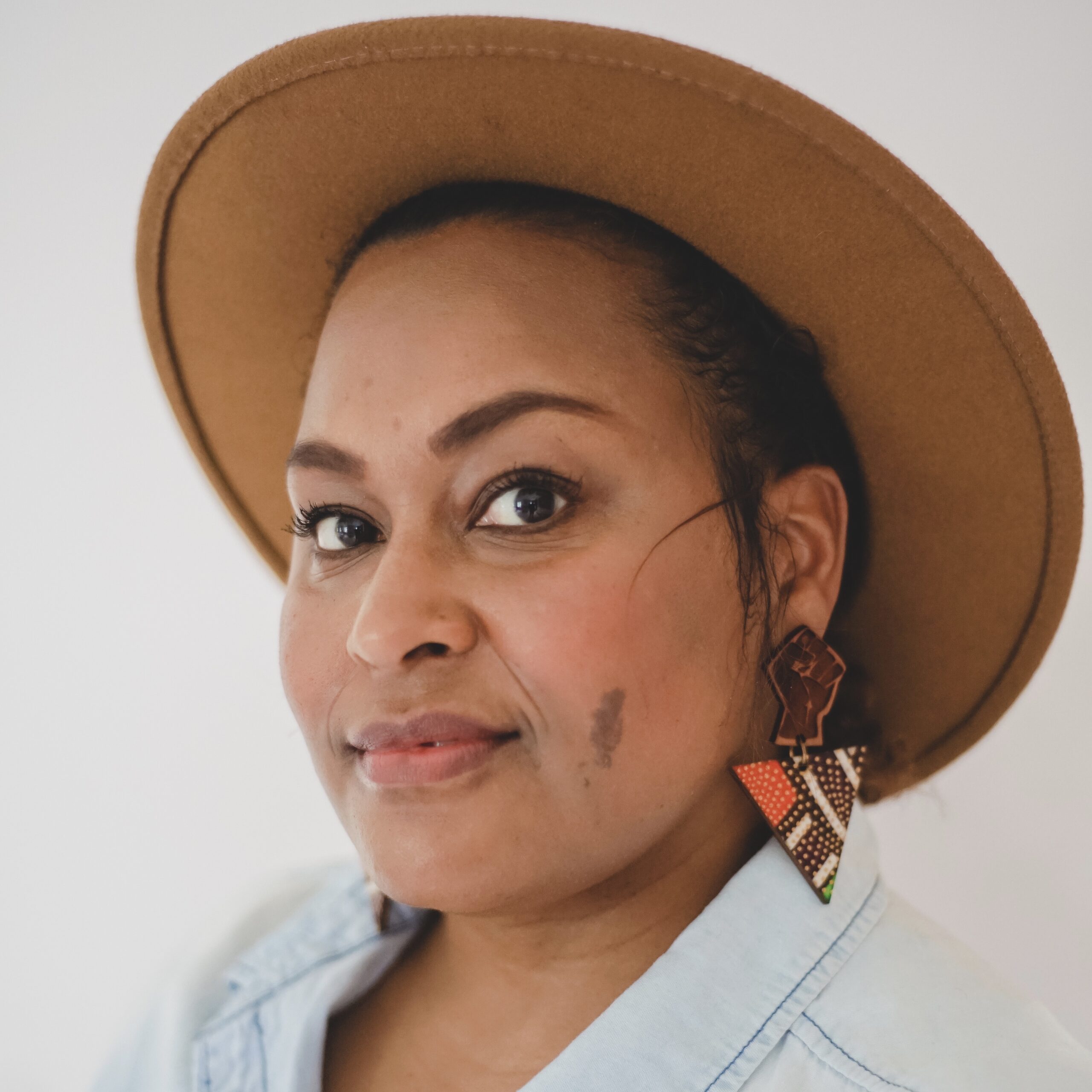 Afro-Latina woman with caramel colored skin smiling at the camera, she is wearing light blue denim shirt, a tan wide brimmed hat and multicolored earrings with a black power fist. 