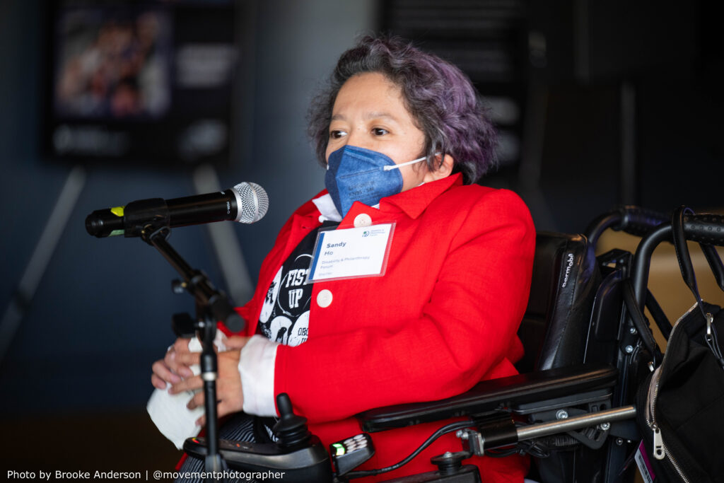 Sandy Ho speaking at the 2025 meeting of the Presidents' Council on Disability Inclusion in Philanthropy. She is sitting in a power wheelchair with a mic in front of her. Sandy is wearing a blue mask, red blazer, white blouse, black graphic T-shirt and black and white pinstripe pants.