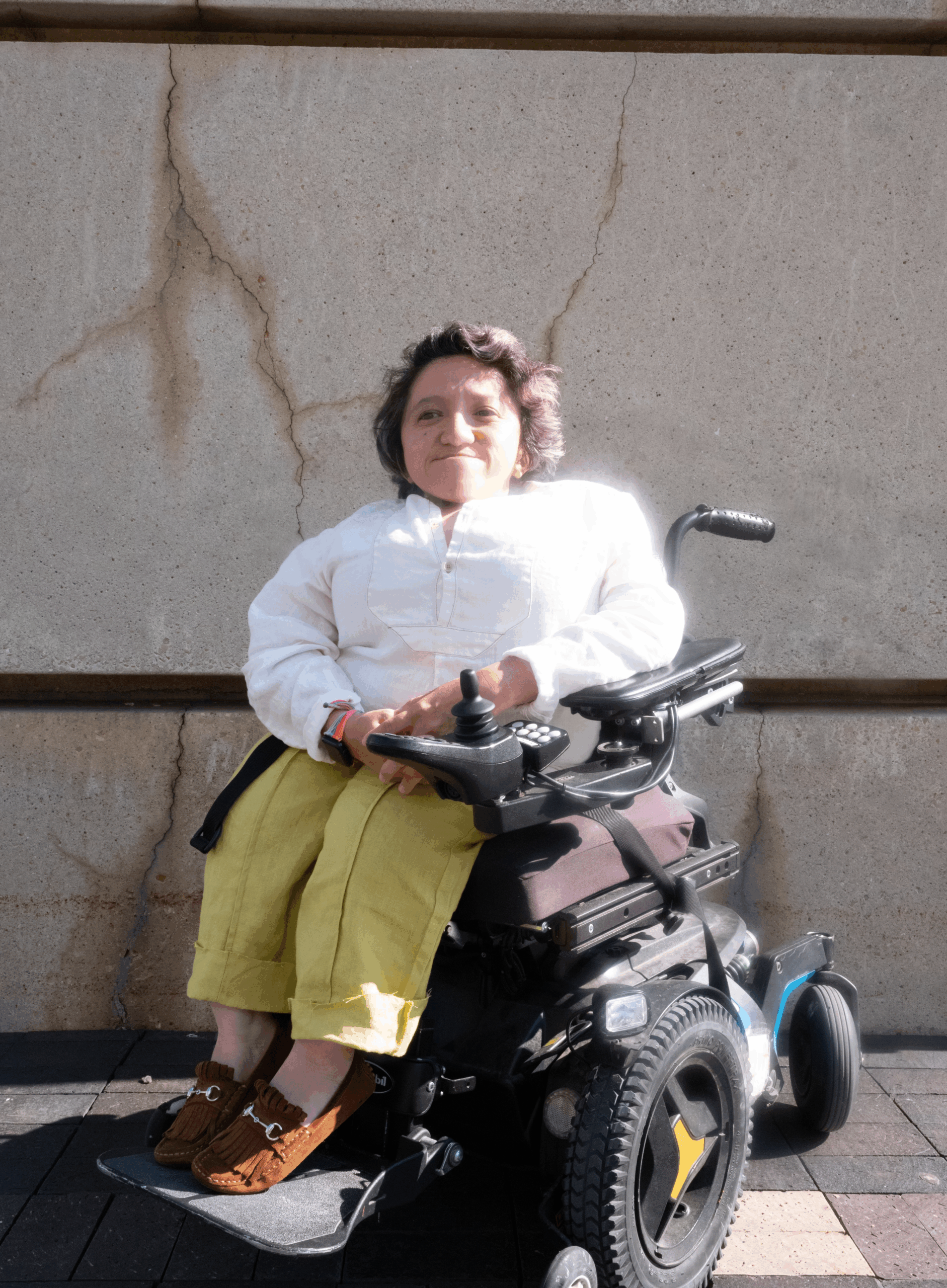Photo taken by Adrian O. Walker. Sandy Ho is sitting outside against a wall in a power wheelchair. She’s wearing a long sleeved white shirt, yellow pants, and brown shoes with a silver buckle. She has short dark wavy hair, and she is smiling at the camera.