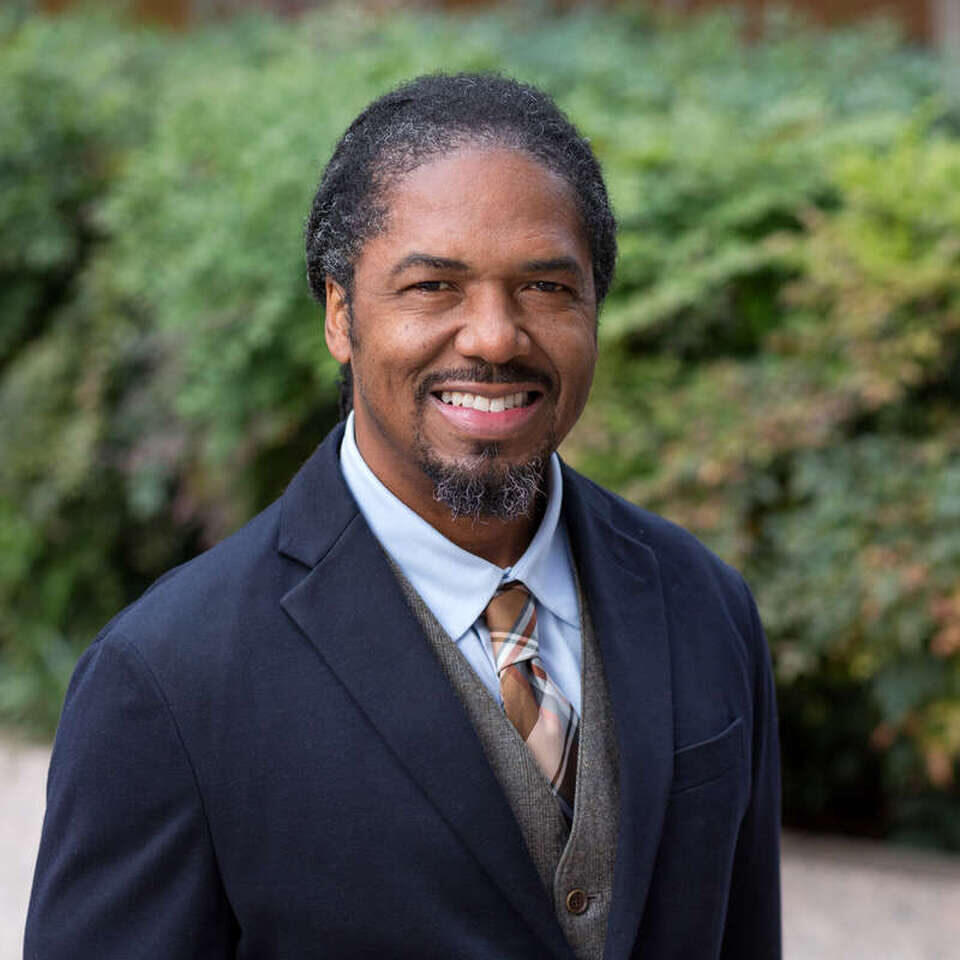Marcus Walton, a Black man with salt-and-pepper hair smiles at the camera. He is wearing a suit with a blue jacket, gray vest, and a light blue shirt. His tie is of several shades of brown, with some red accent colors.