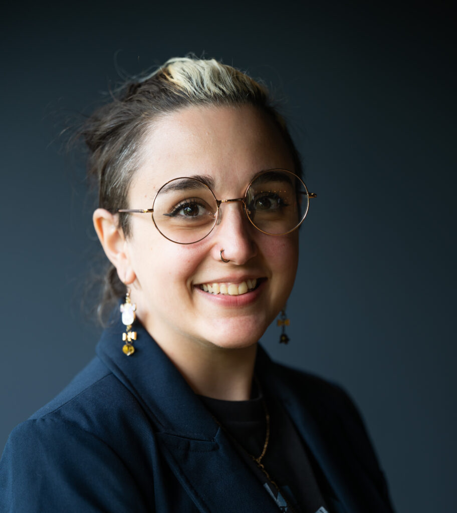 Noor is a lightly tanned Palestinian woman. She is pictured smiling against a navy blue background and looking directly at the camera. Her brown hair is pulled into a ponytail with a strip of white down the center. She is wearing gold rimmed circular glasses, dangling cow earrings with a bell attached, and a navy blue blazer.