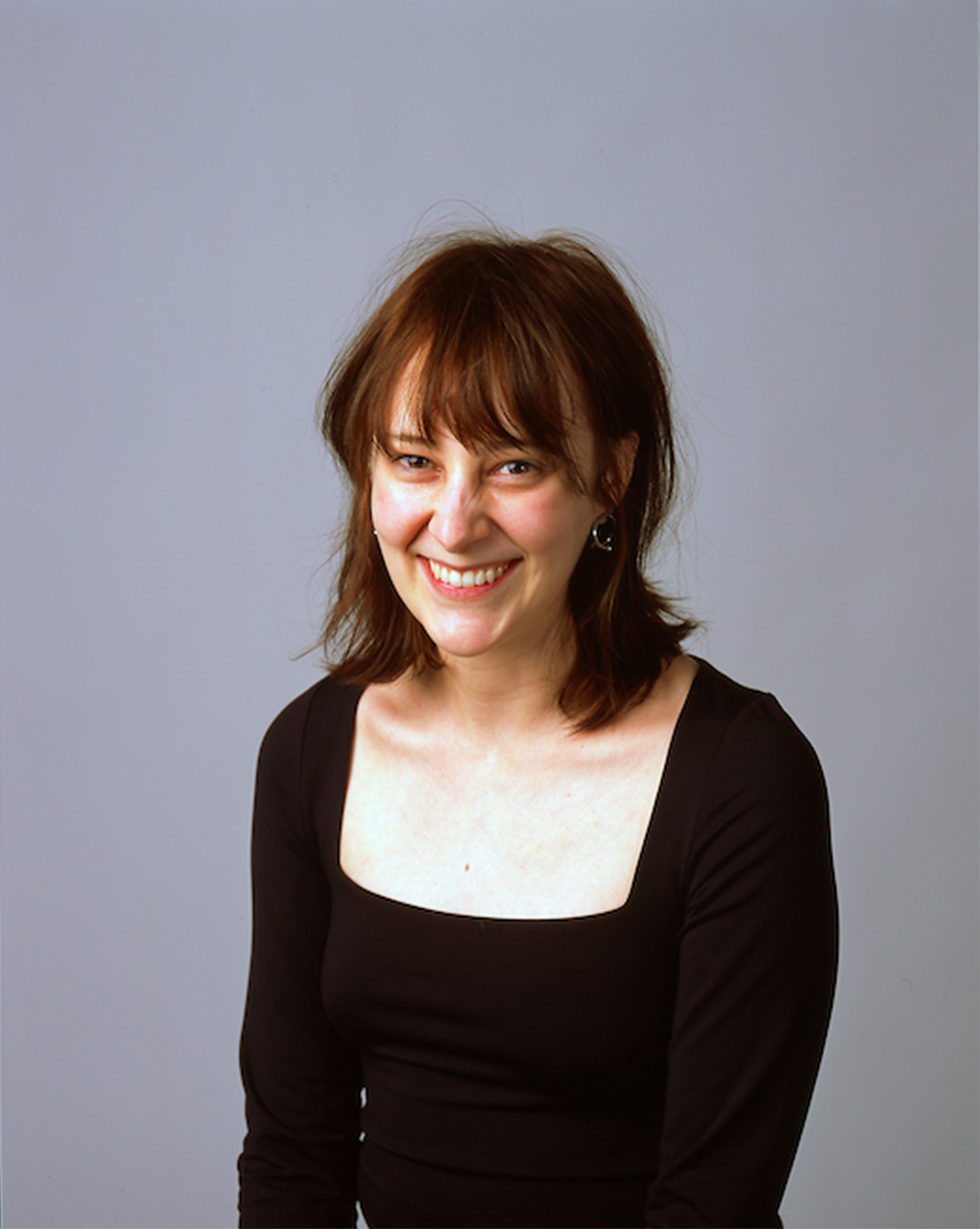 Olivia sits against a plain light grey background. She is a white woman in her early thirties with shoulder-length brown hair and bangs, smiling warmly at the camera. She is wearing a fitted black top with a square neckline.