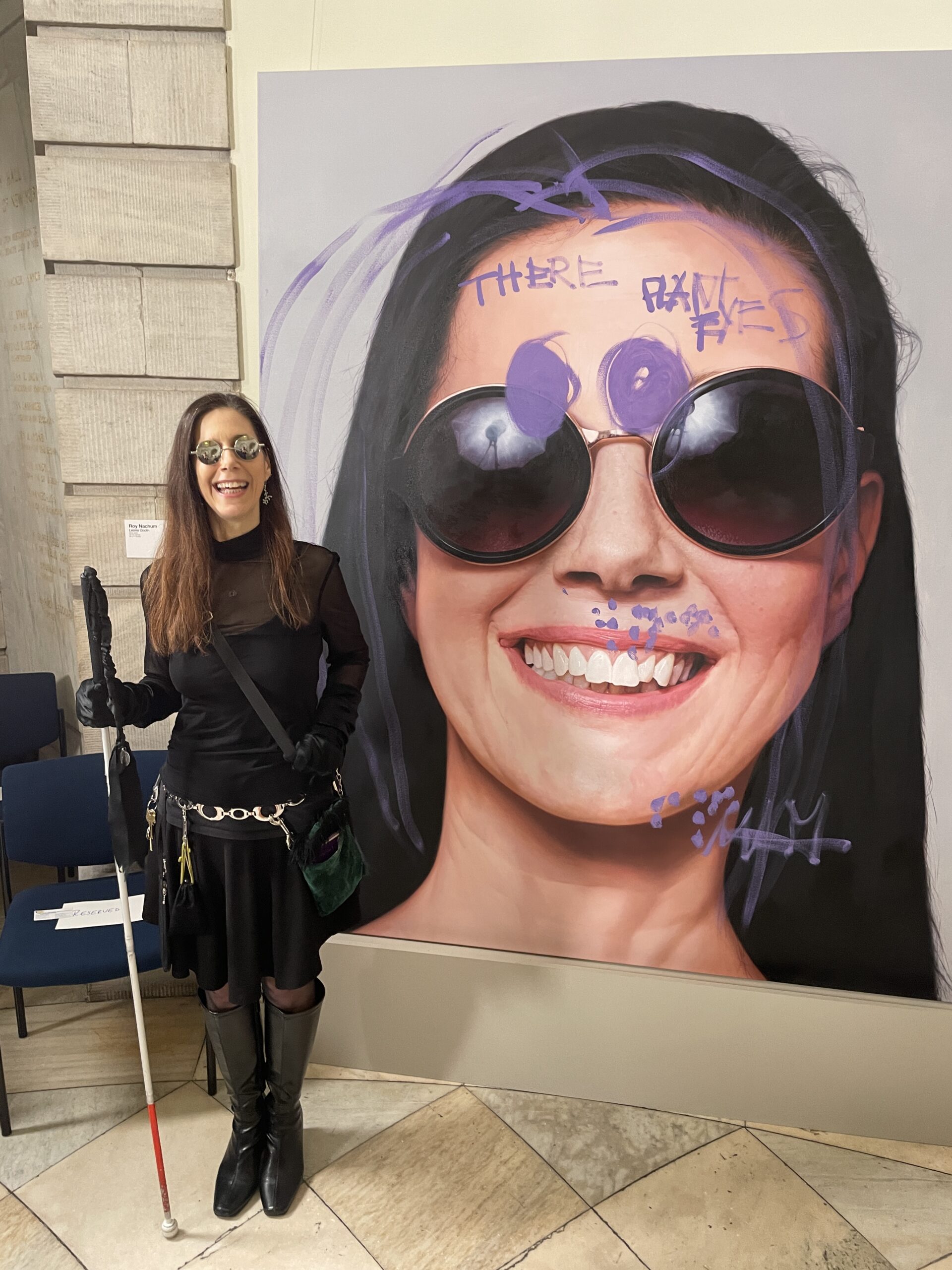 Leona stands next to a large, hyper-realistic painting of her face that has been graffitied by her with ultraviolet paint--—strokes of hair, circles for sunglass lenses, random dots, “There Plant Eyes” on her forehead. She’s wearing black gloves, sheer black turtleneck, round mirror sunglasses, Moses (her white cane) is in her hand, and she’s smiling.