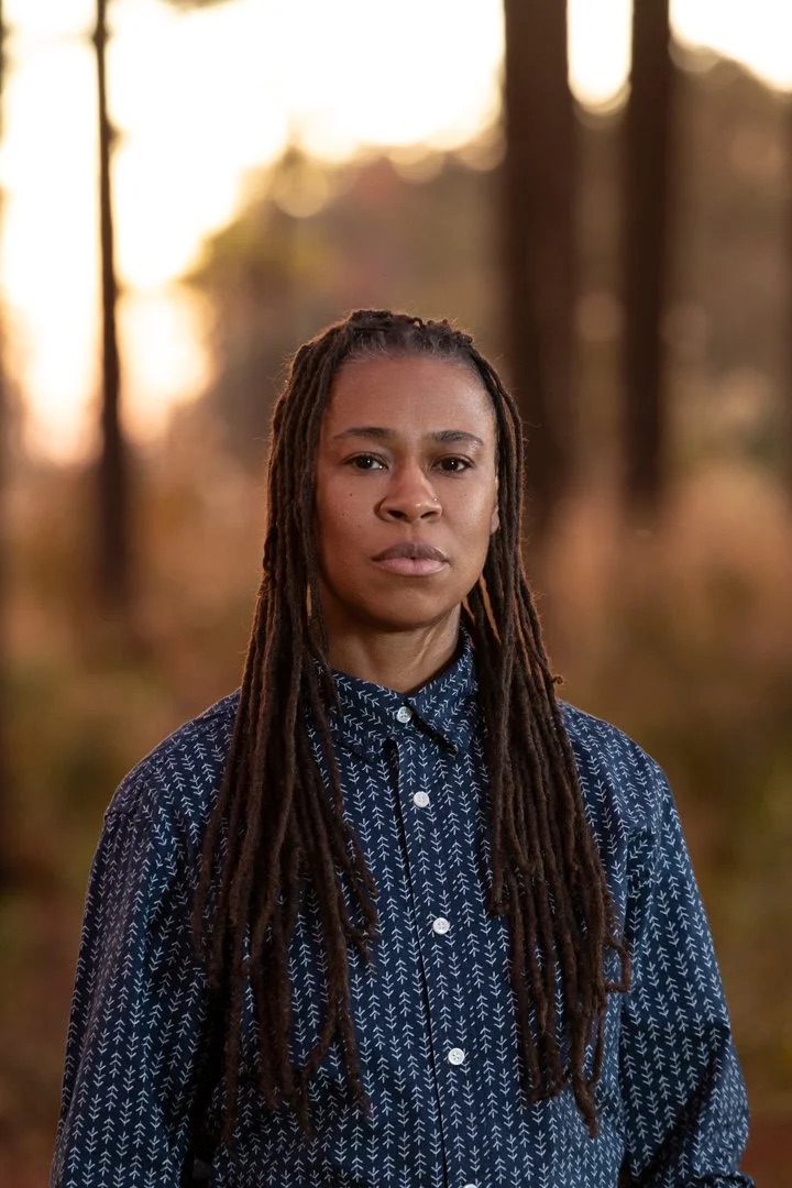 A Black masculine presenting person wearing a button down shirt looking at the camera. They are standing outside in what appears to be a wooded area.