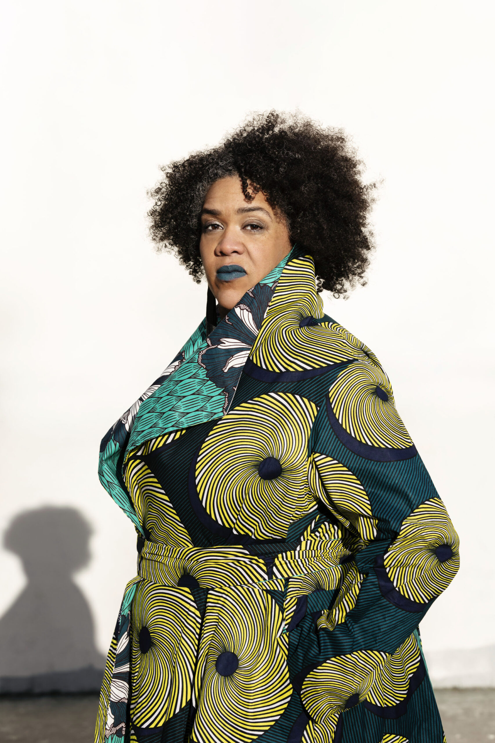 Brown skinned black woman with a big curly fro and blue lipstick looking ready at the camera with hands in her pockets and the collar popping on a blue, yellow and white swirling patterned long draping coat.