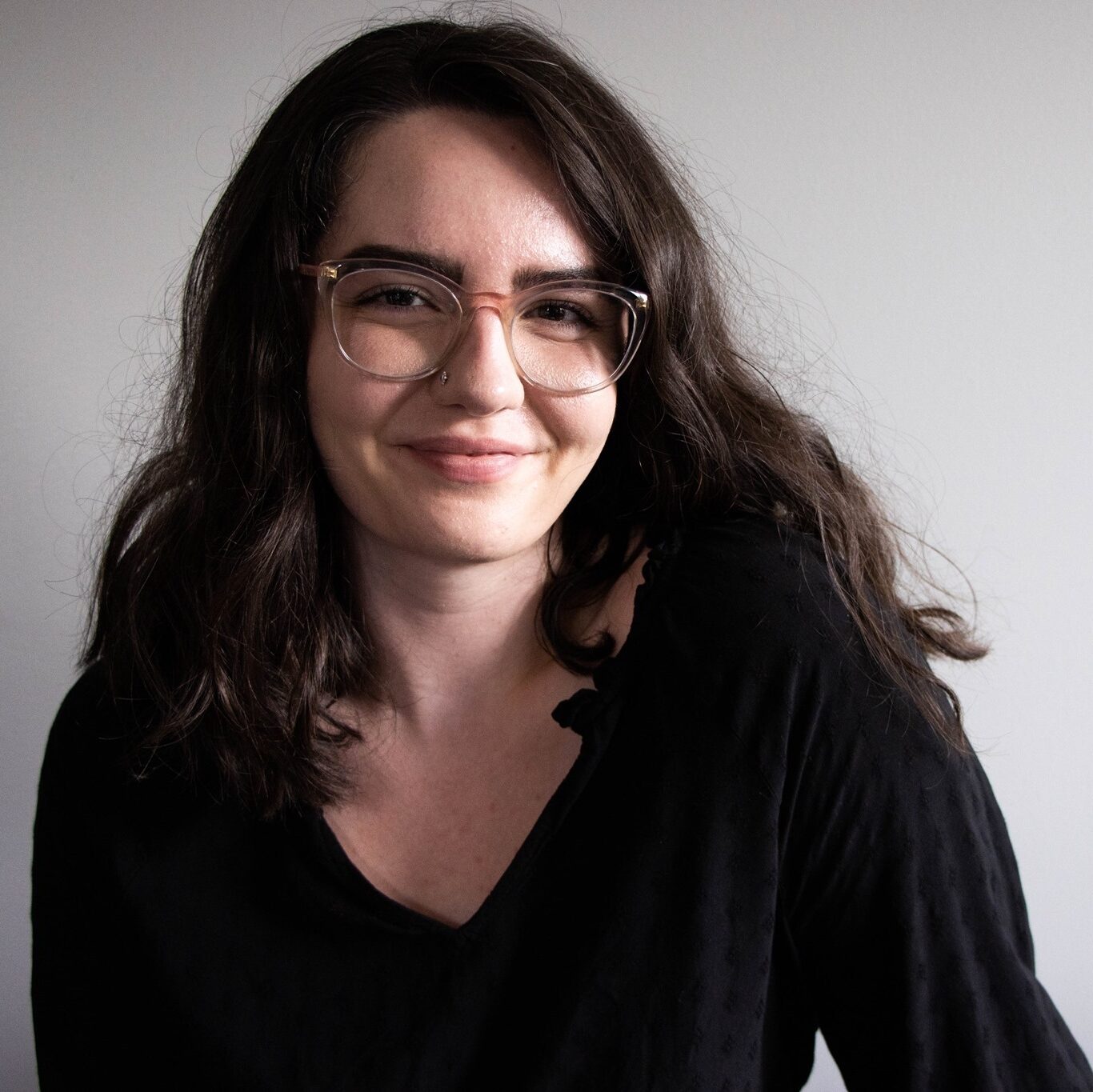 A headshot of a white woman with brown wavy hair just beyond her shoulders. The background is light gray. She is wearing clear framed glasses, a nose ring stud, and has on a black blouse. She is smiling.