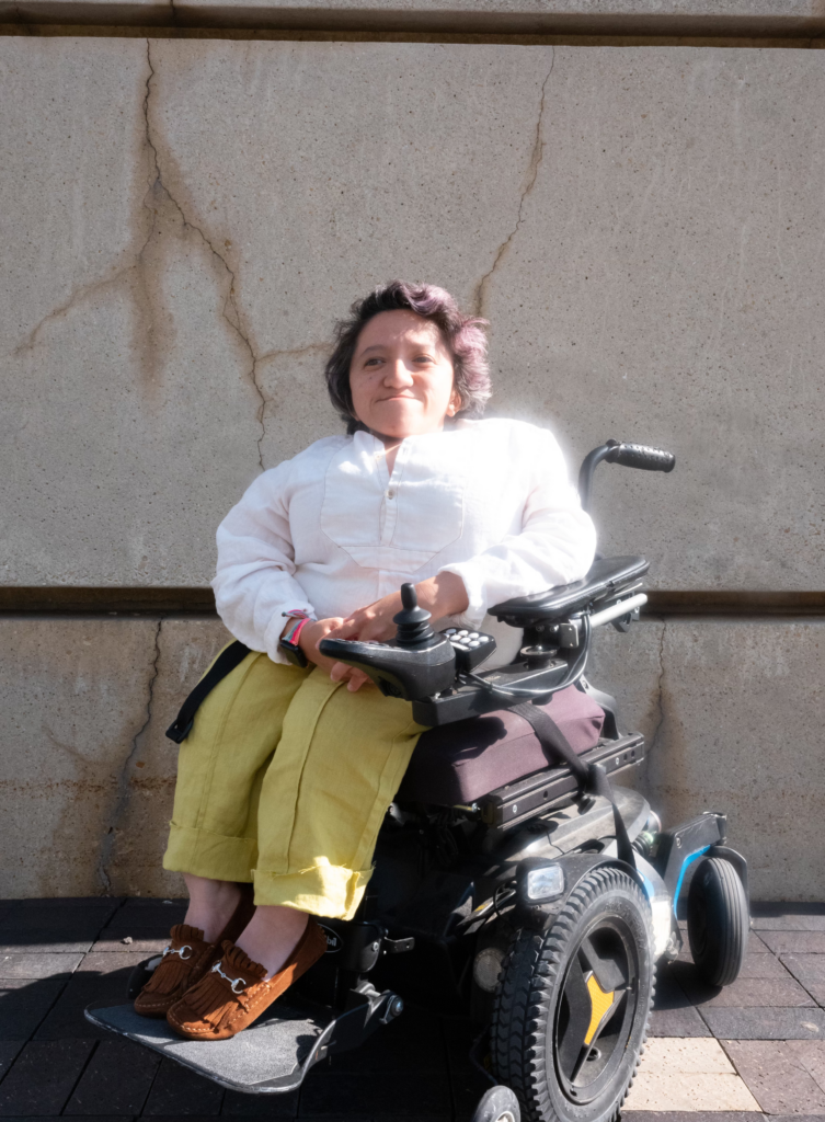 photo of sandy ho against a cement wall with visible cracks. she is sitting in a black power wheelchair wearing a white button up shirt, yellow pants, and brown shoes. she has short dark wavy hair, and is smiling off to the side.